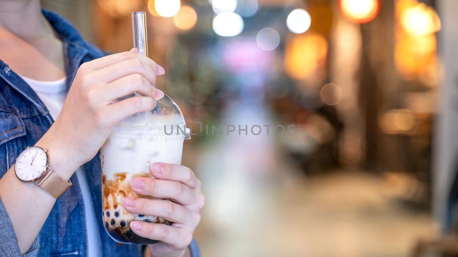 Young girl in denim jacket is drinking brown sugar flavored tapioca pearl bubble milk tea with glass straw in night market of Taiwan, close up, bokeh