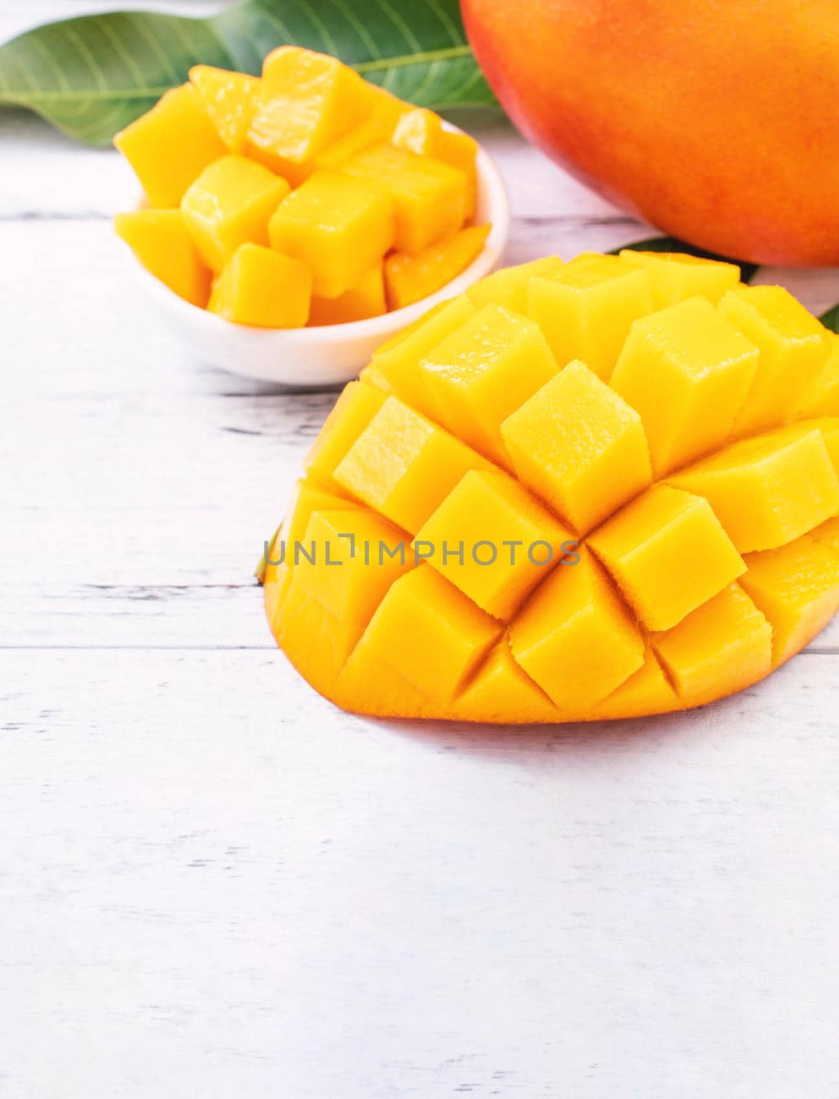 Beautiful chopped mango chunks with green leaves on bright white color timber background. Copy space, close up, macro. Tropical fruit concept.