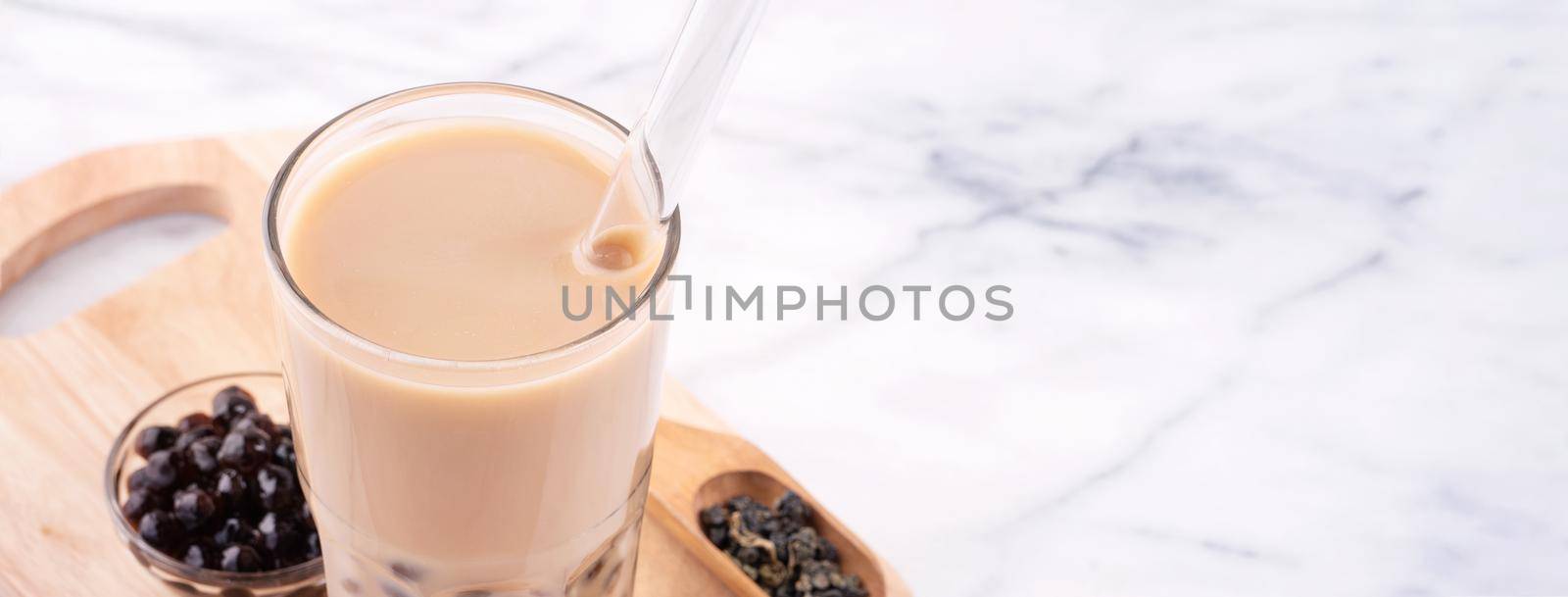 Tapioca pearl ball bubble milk tea, popular Taiwan drink, in drinking glass with straw on marble white table and wooden tray, close up, copy space.