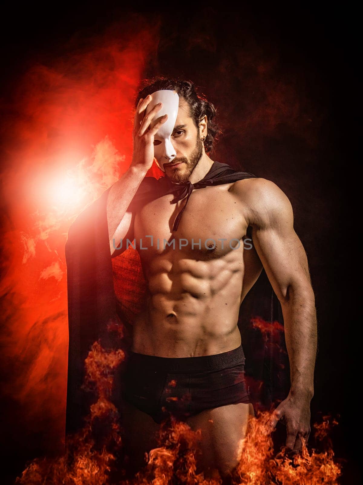 Shirtless muscular male with naked torso in cloak and underpants standing in darkness, among fire and covering part of face with white mask