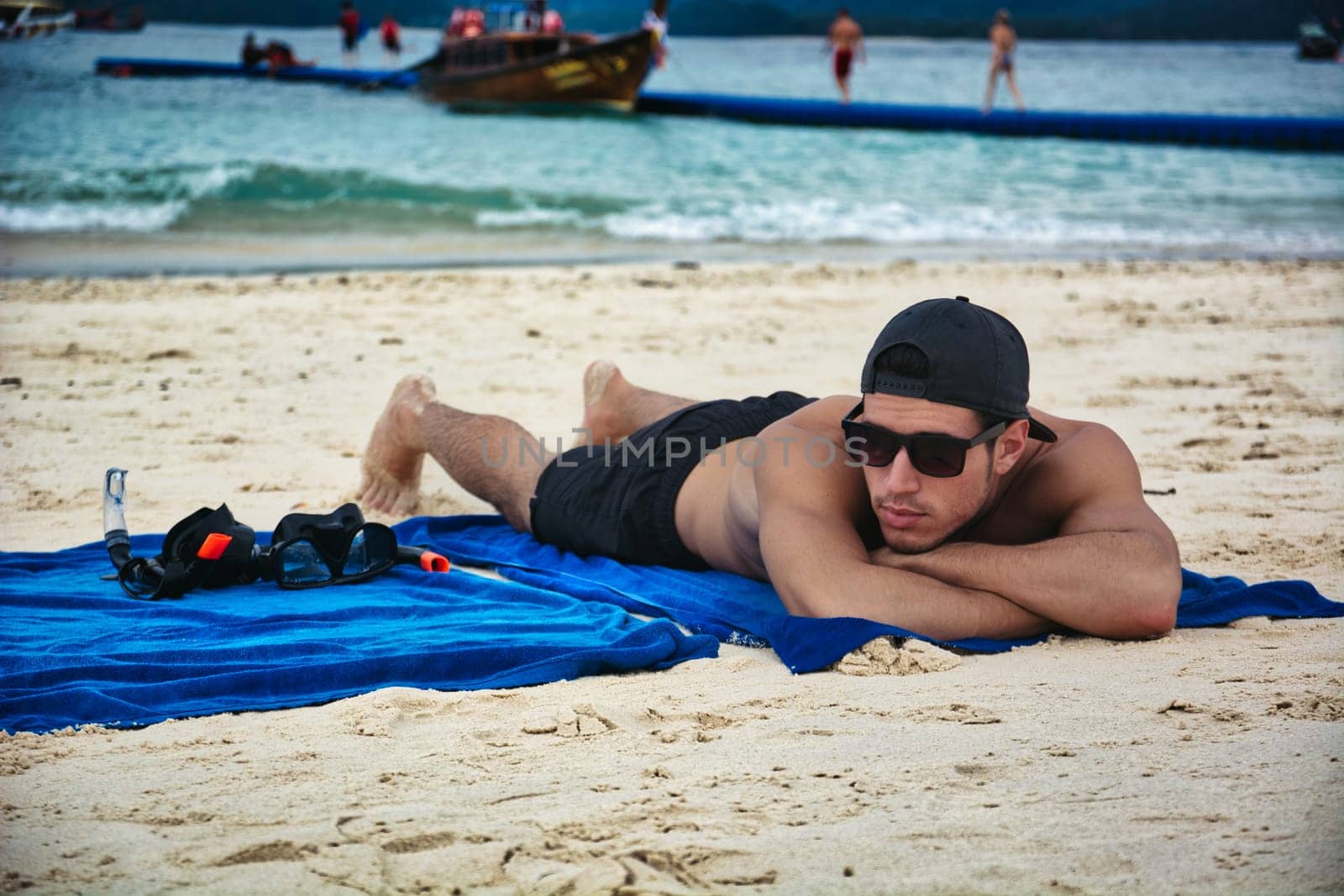 Full body shot of a handsome young man laying on a tropical beach in Phuket Island, Thailand, on a beach towel, shirtless wearing baseball cap and boxer shorts, showing muscular fit body