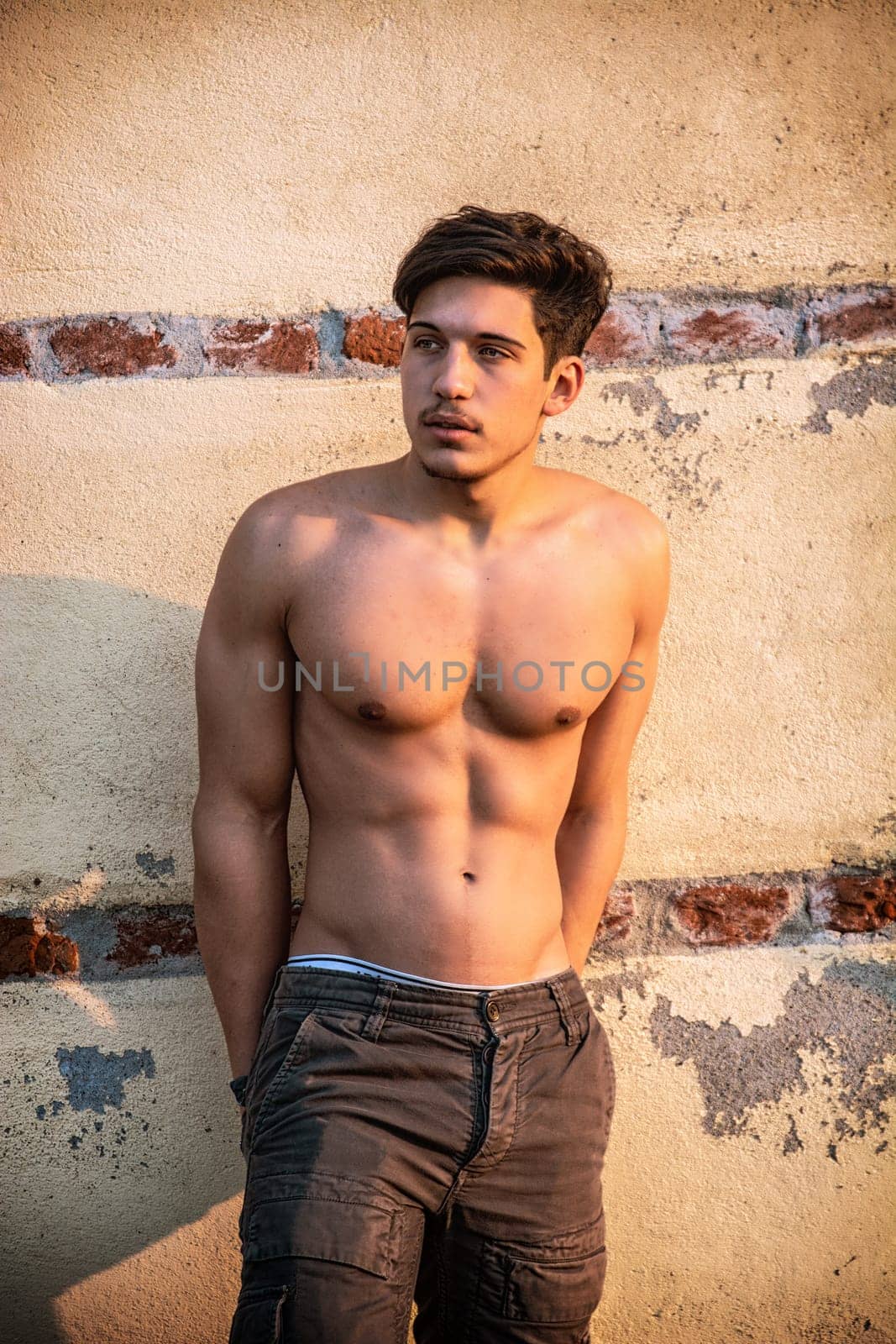 Athletic young man shirtless in jeans in front of concrete wall outside, with arms crossed on his chest, showing sexy body, looking away to a side