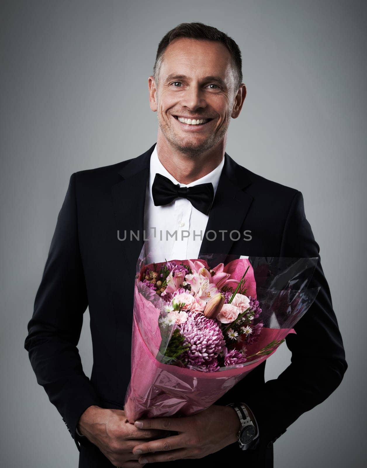 Portrait, man and bouquet of flowers, suit and happiness for Valentines day, romance and on grey studio background. Face, male and guy with floral gift, smile and gesture for love, romantic and happy by YuriArcurs