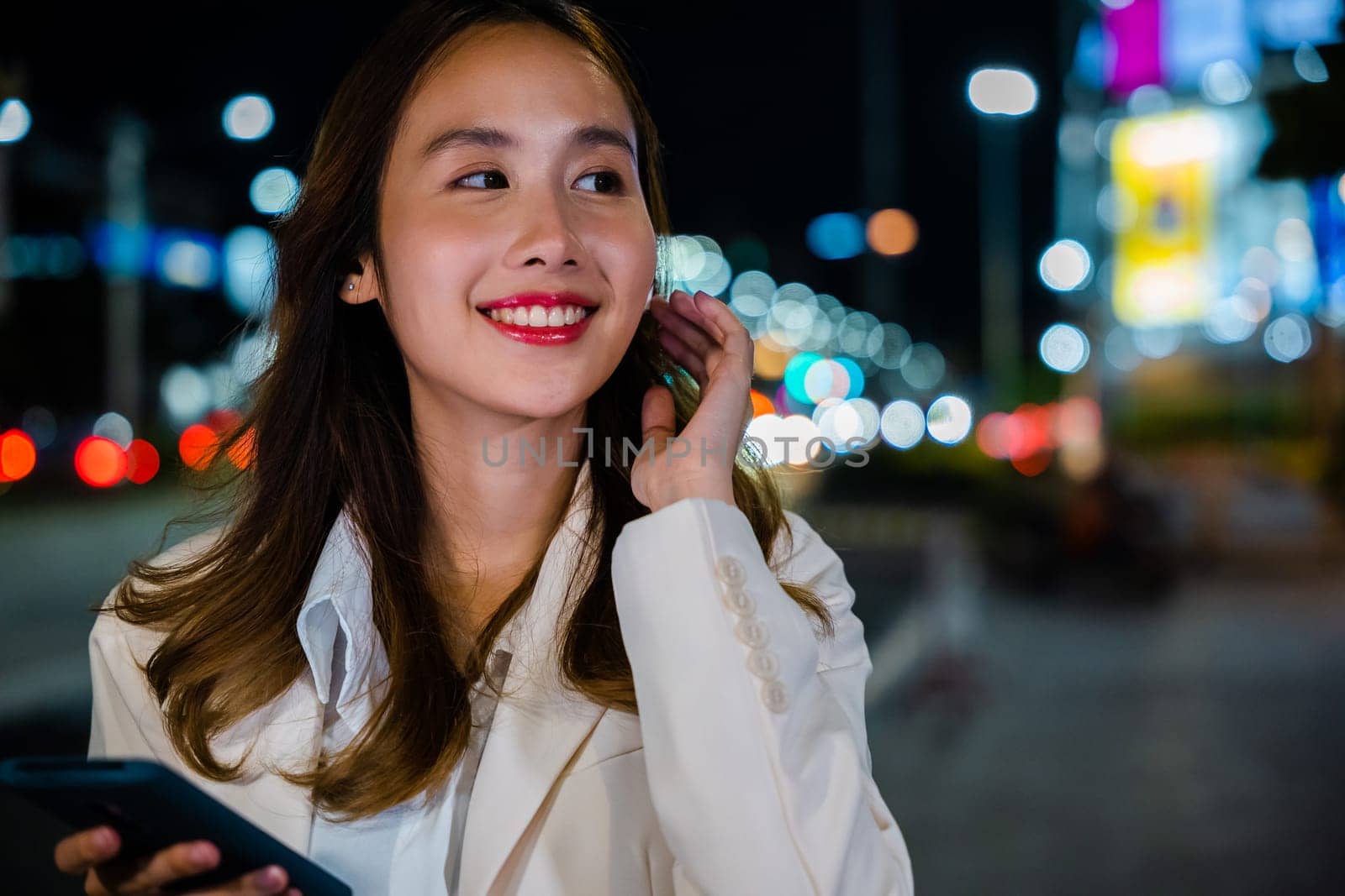 Smiling woman enjoying music through earphones attached to her mobile phone while strolling the night city streets. Wireless technology and digital devices make life convenient. Portrait of happy girl
