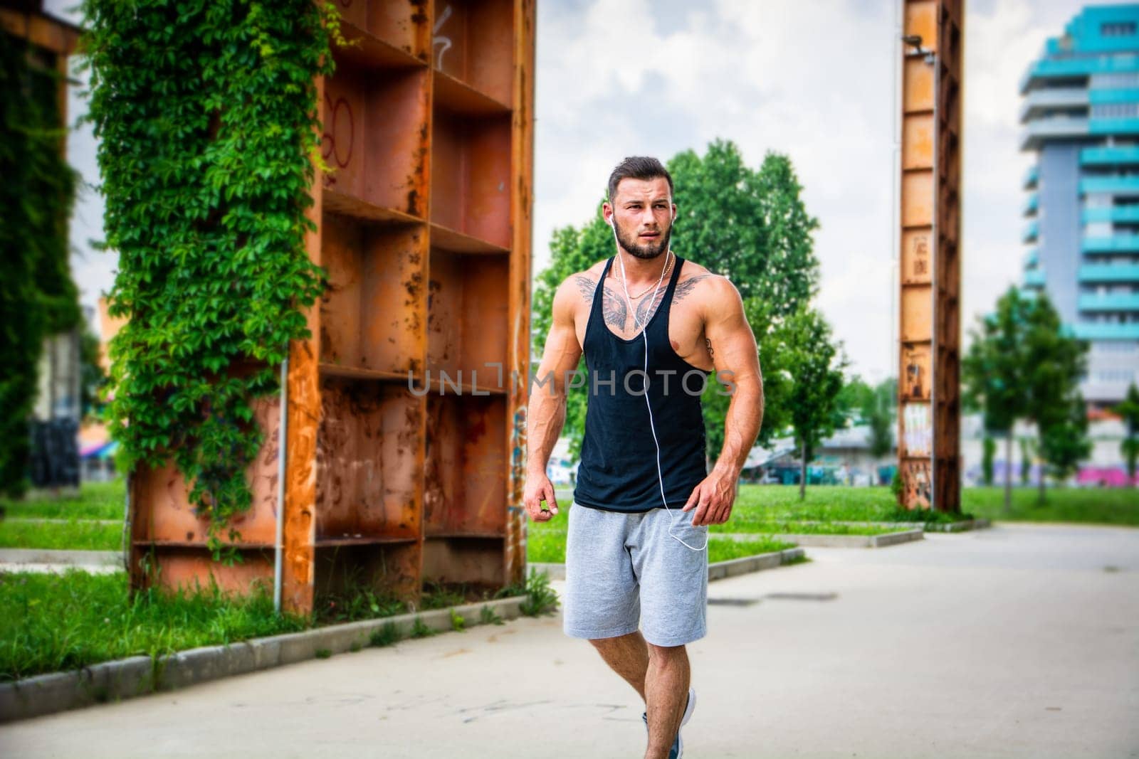 Handsome muscular athletic man running and jogging in city park during the day, wearing black tank top