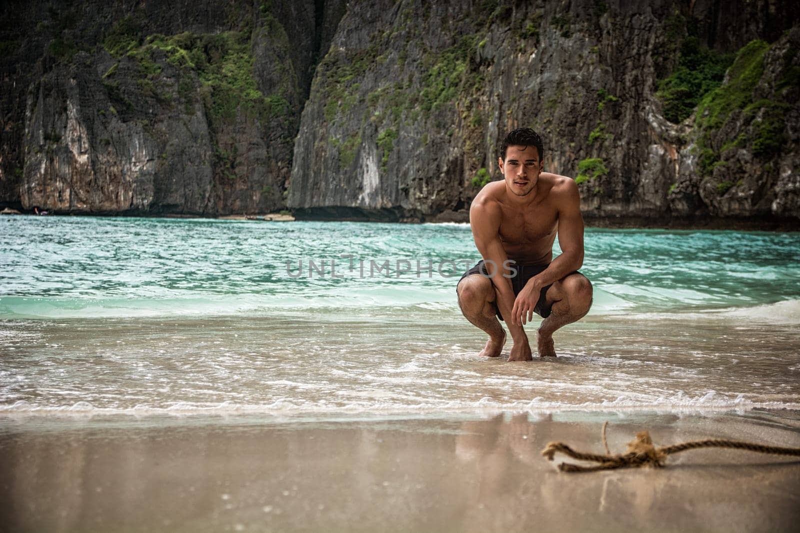 Attractive young man crouching on a beach in Phuket Island, Thailand, shirtless wearing boxer shorts, showing muscular fit body, looking at camera