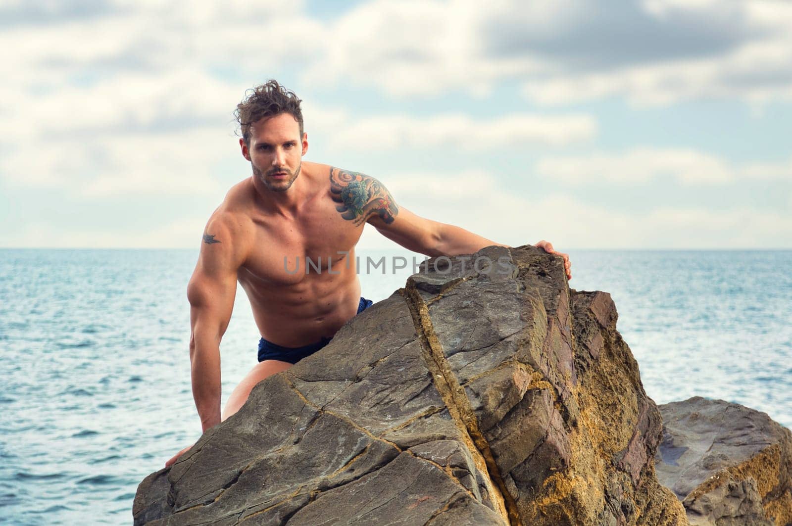Attractive muscular shirtless athletic man standing on rock next to water by sea or ocean shore, looking at camera in a cloudy summer day