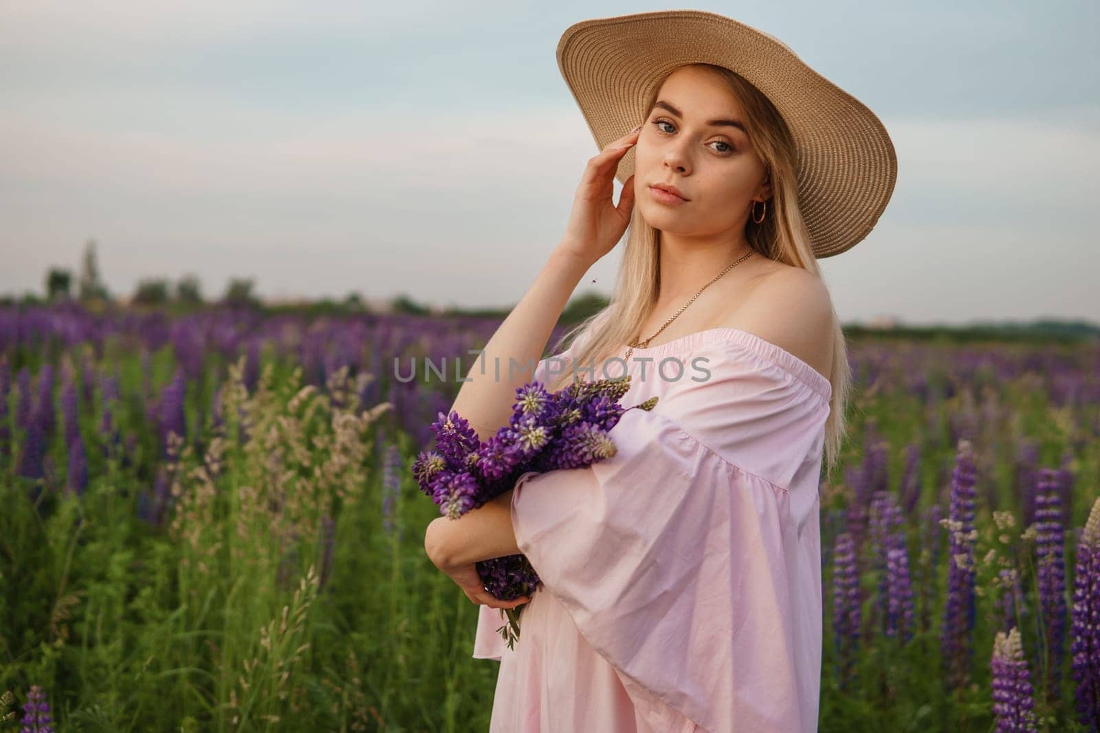 A beautiful woman in a straw hat walks in a field with purple flowers. A walk in nature in the lupin field.