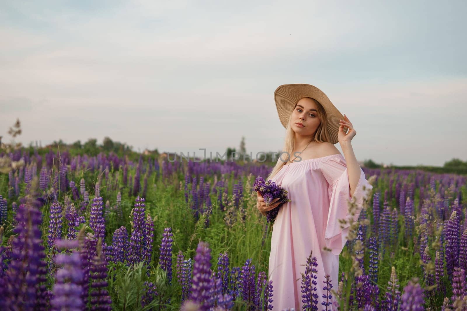 A beautiful woman in a straw hat walks in a field with purple flowers. A walk in nature in the lupin field.