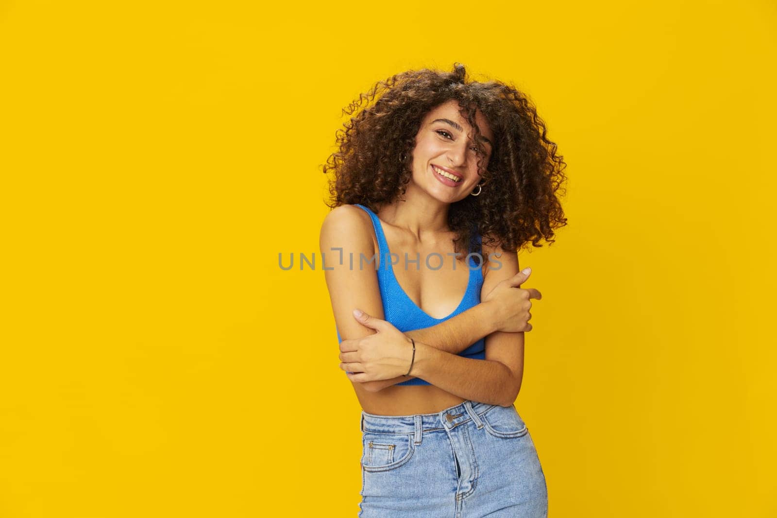 Woman with curly afro hair in a blue t-shirt on. yellow background signs with her hands, look into the camera, smile with teeth and happiness, copy space. High quality photo