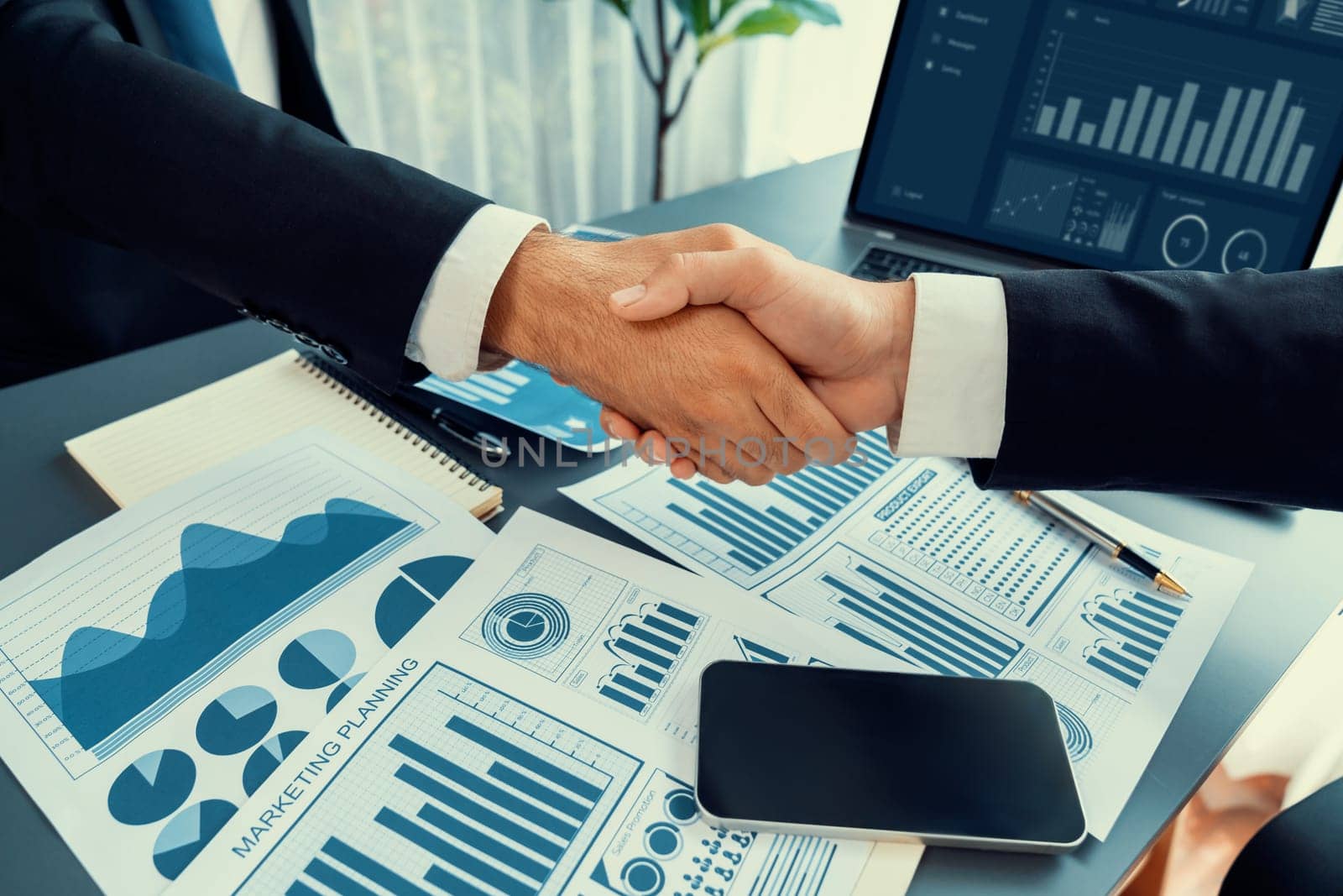 Closeup professional businessman shaking hands over desk in modern office after successfully analyzing pile of dashboard data paper as teamwork and integrity handshake in workplace concept. fervent