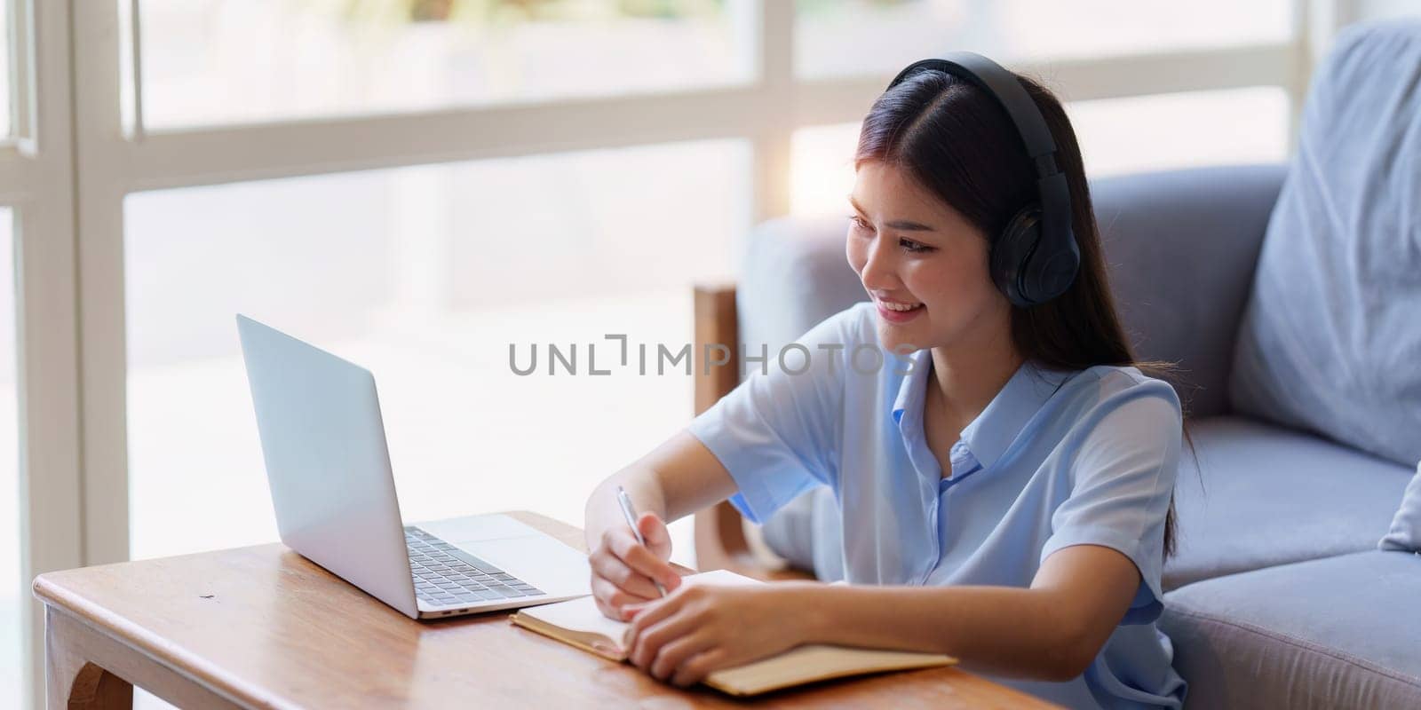 Online education, e-learning. Asian woman in stylish casual clothes, studying using a laptop, listening to online lecture, taking notes, online study at home.