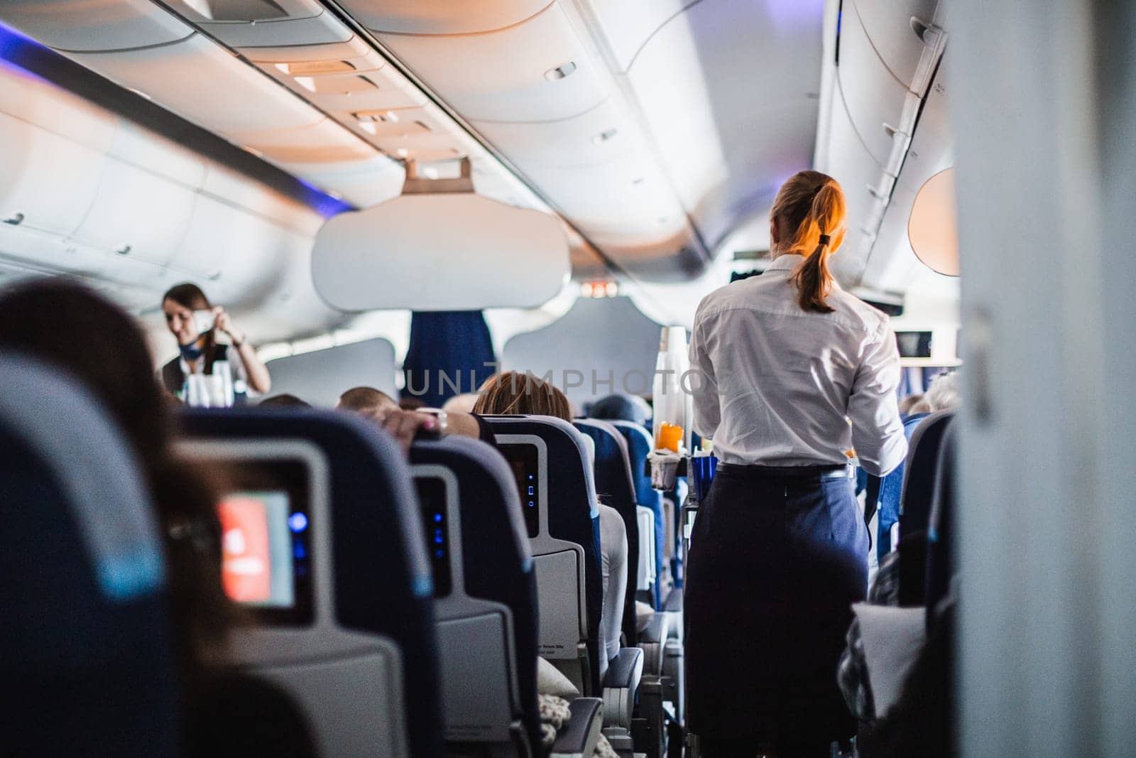 Interior of airplane with passengers on seats and stewardess in uniform walking the aisle, serving people. Commercial economy flight service concept