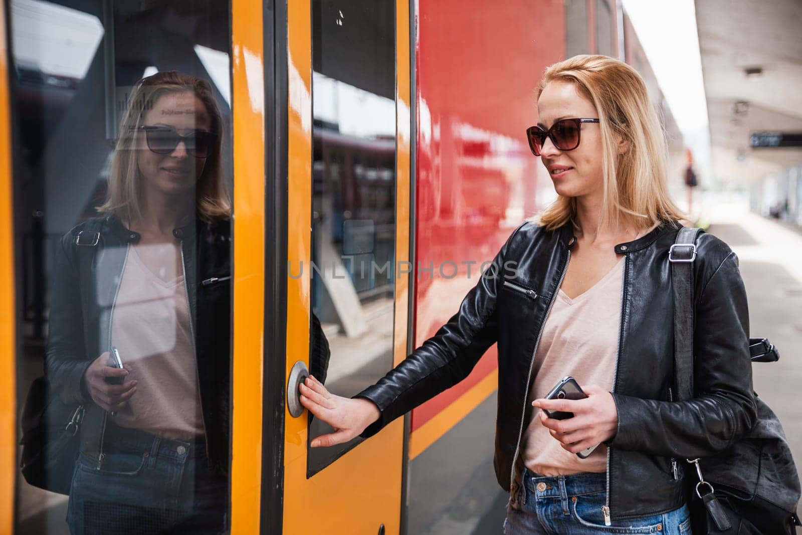 Young blond woman in jeans, shirt and leather jacket wearing bag and sunglass, presses door button of modern speed train to board on train station platform. Travel and transportation.