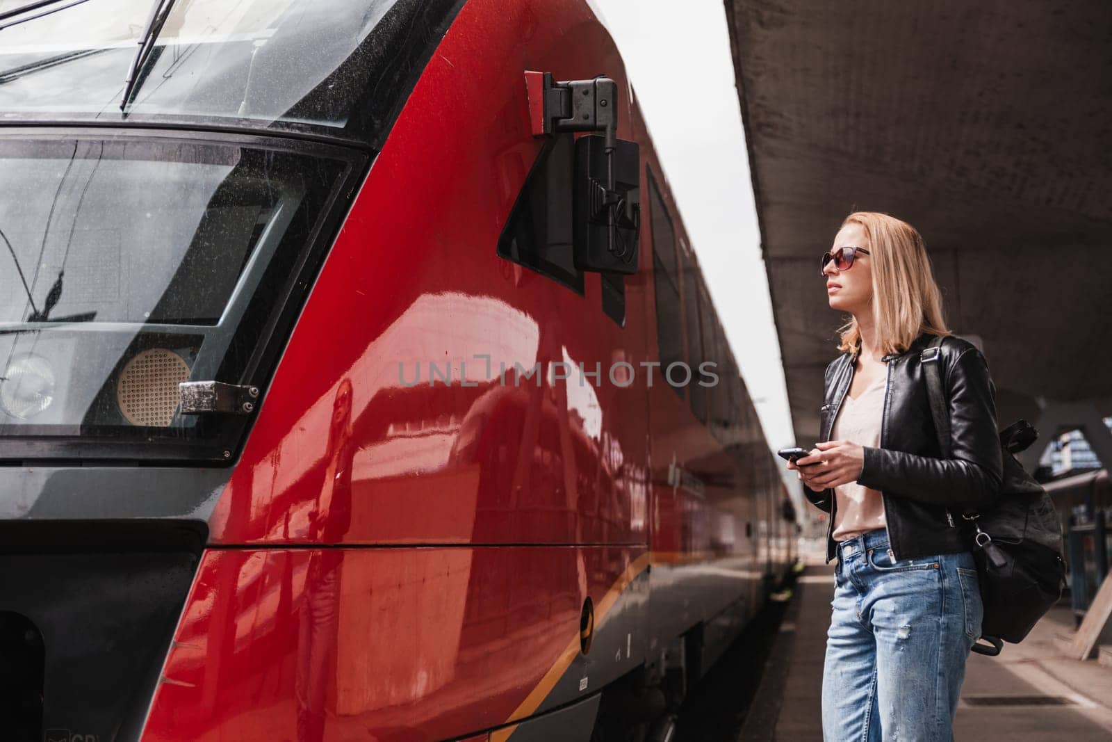 Young blond woman in jeans, shirt and leather jacket wearing bag and sunglass, embarking red modern speed train on train station platform. Travel and transportation