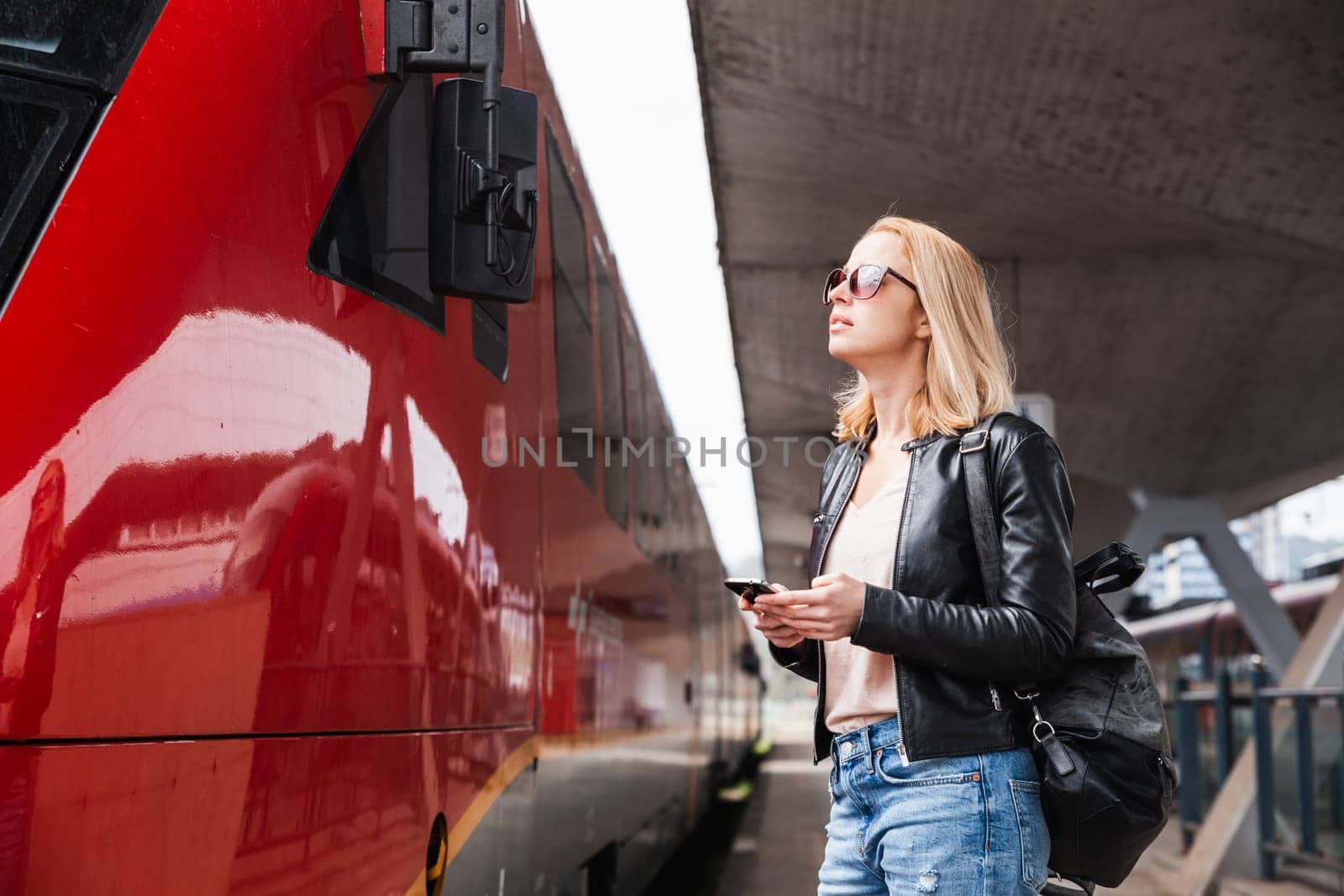 Young blond woman in jeans, shirt and leather jacket wearing bag and sunglass, embarking red modern speed train on train station platform. Travel and transportation. by kasto