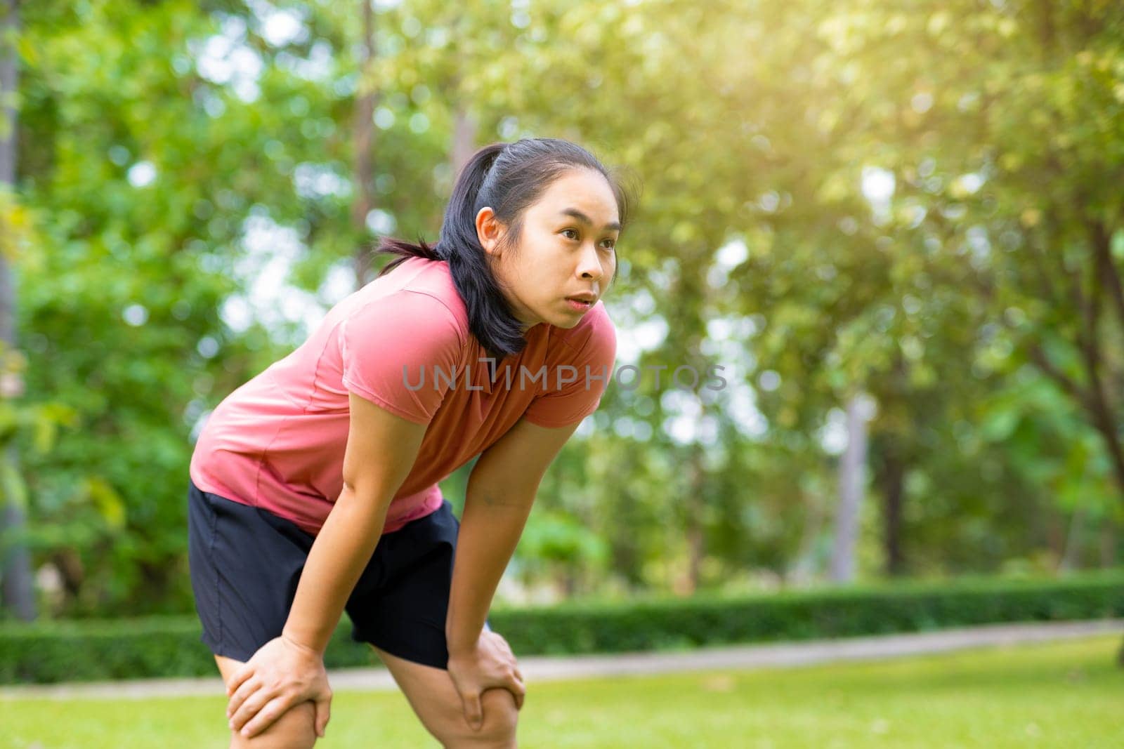Young Asian woman standing bent over and breathing after running in the park. Young healthy woman taking a break after running. Healthy lifestyle concept.