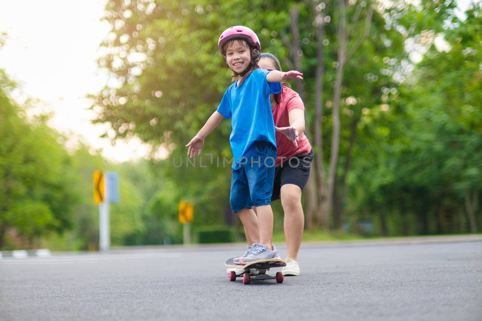 Active little girl and mom enjoy skateboarding. Cute little girl wearing helmet practicing skateboarding in park. Mother trains her daughter to skateboard. Outdoor sports for children. by TEERASAK