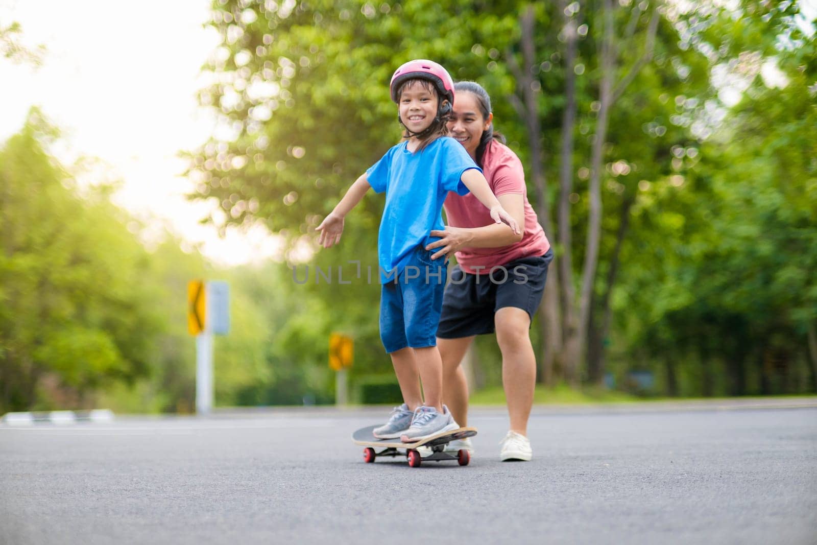 Active little girl and mom enjoy skateboarding. Cute little girl wearing helmet practicing skateboarding in park. Mother trains her daughter to skateboard. Outdoor sports for children.