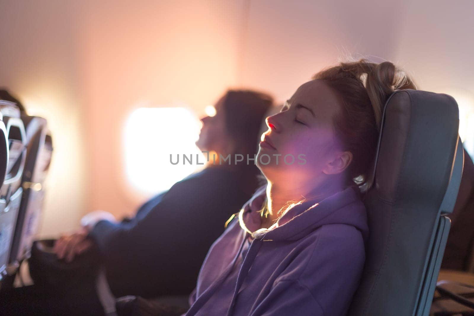 Out of fokus image of tired blonde casual caucasian woman wearing sporty hoodie napping on seat while traveling by airplane. Commercial transportation by planes. Authentic image of real people. by kasto