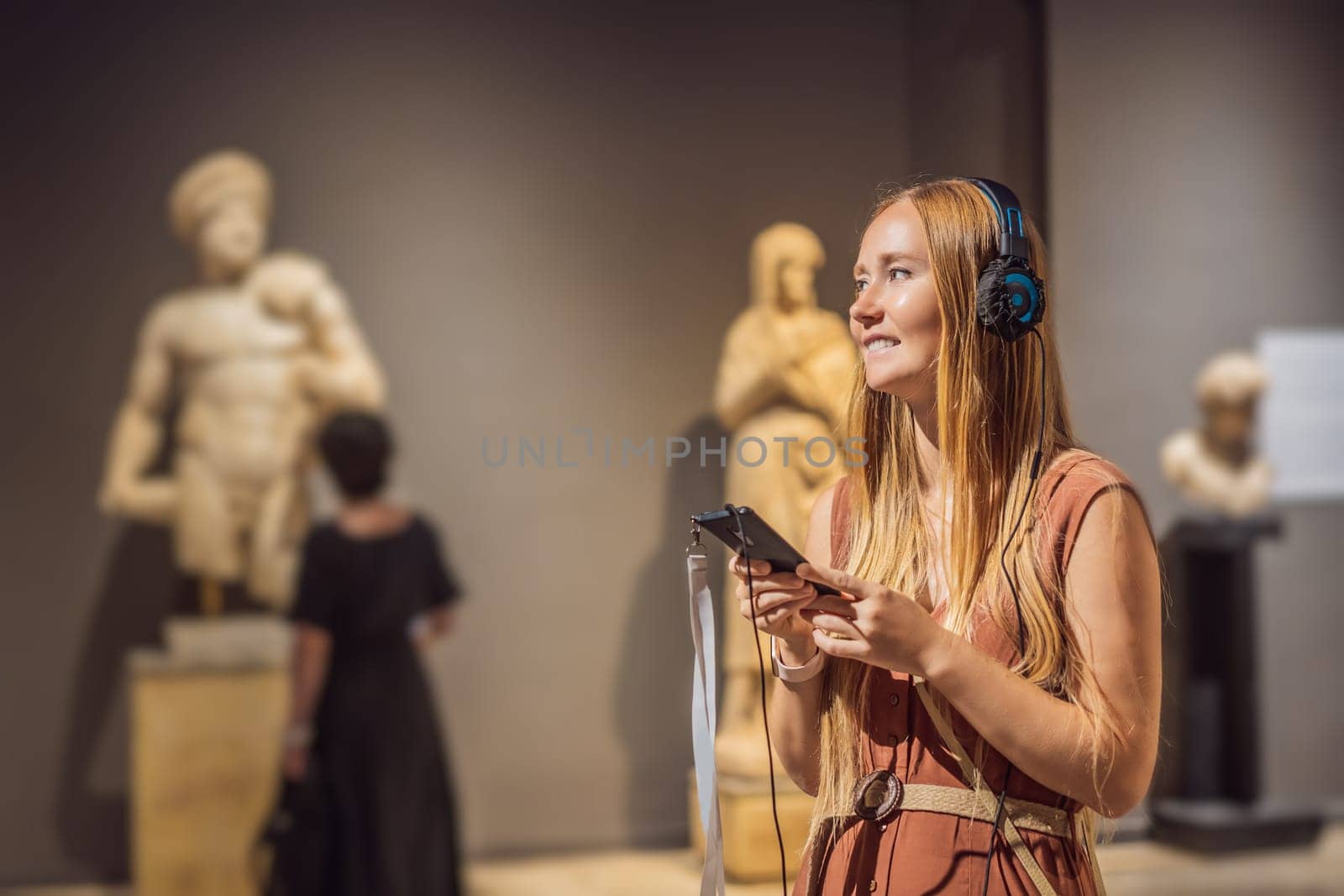 Portrait of contemporary young woman looking at sculptures and listening to audio guide at museum exhibition.