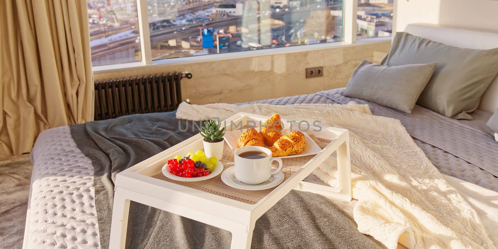 White Tray with tasty breakfast on bed in light bedroom with big windows in skyscraper. white table with tasty breakfast in bed at bed room in skyscraper