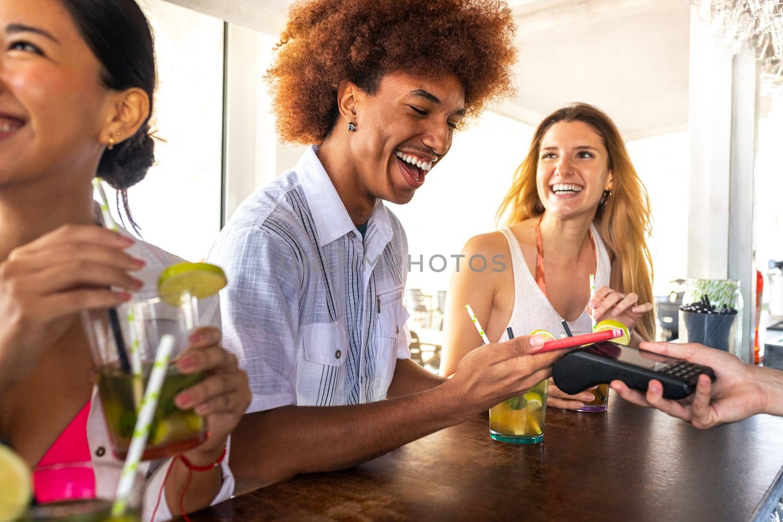 African American man paying bill at beach bar with phone. Young black male paying round of cocktail drinks with mobile phone on summer vacation. Technology and leisure concept.