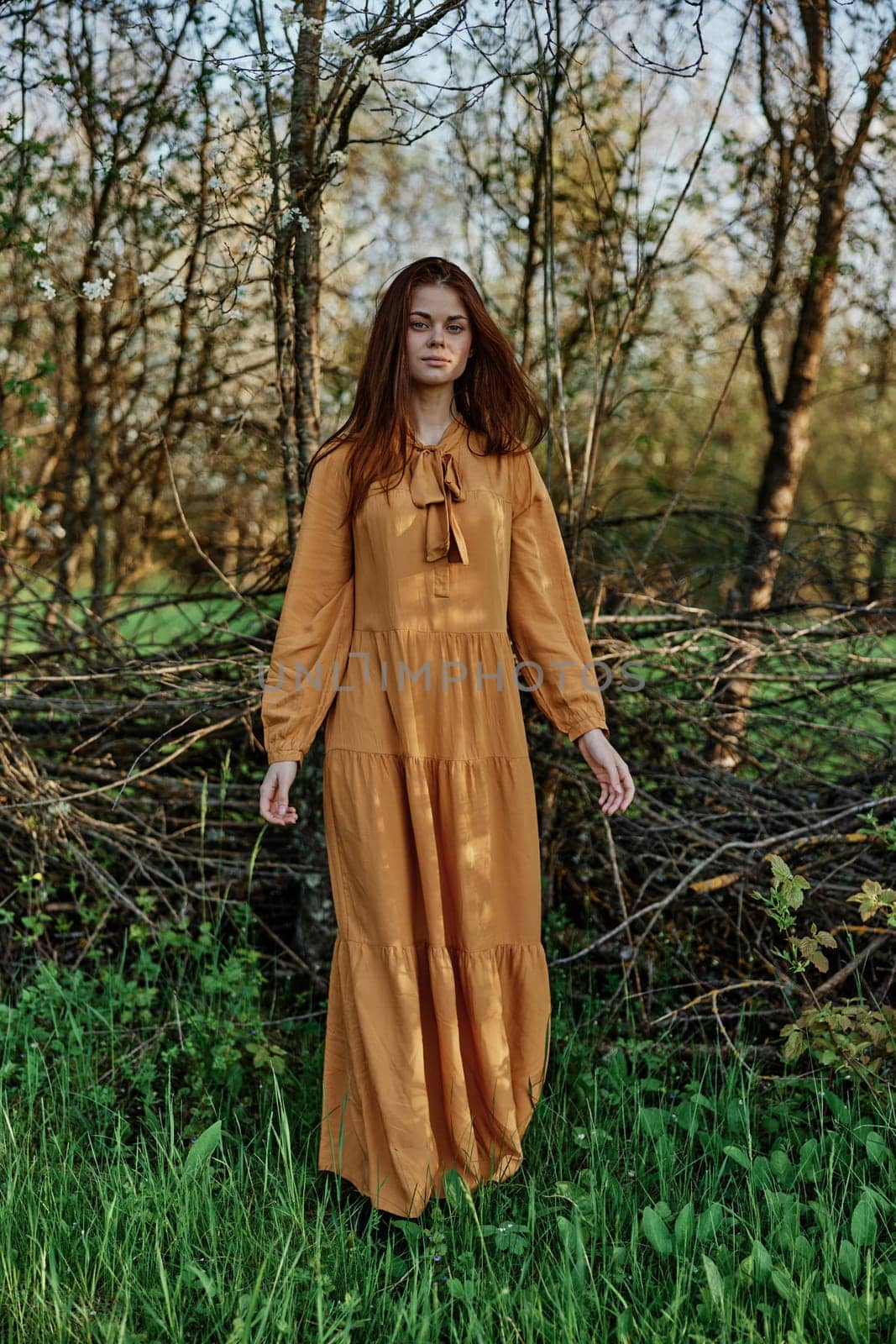 vertical photo of a beautiful, elegant woman in a stylish orange dress standing in the countryside in the shade of trees. High quality photo