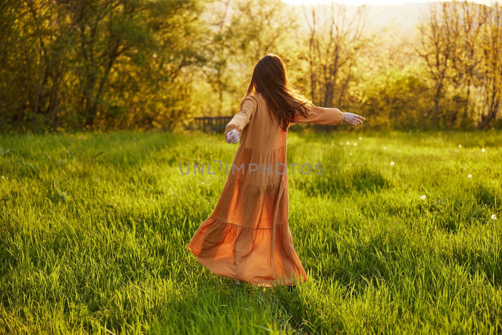 a relaxed slender woman enjoys the sunset standing in a green field with tall grass in an orange dress with her back to the camera, in warm summer weather. Horizontal photo. High quality photo