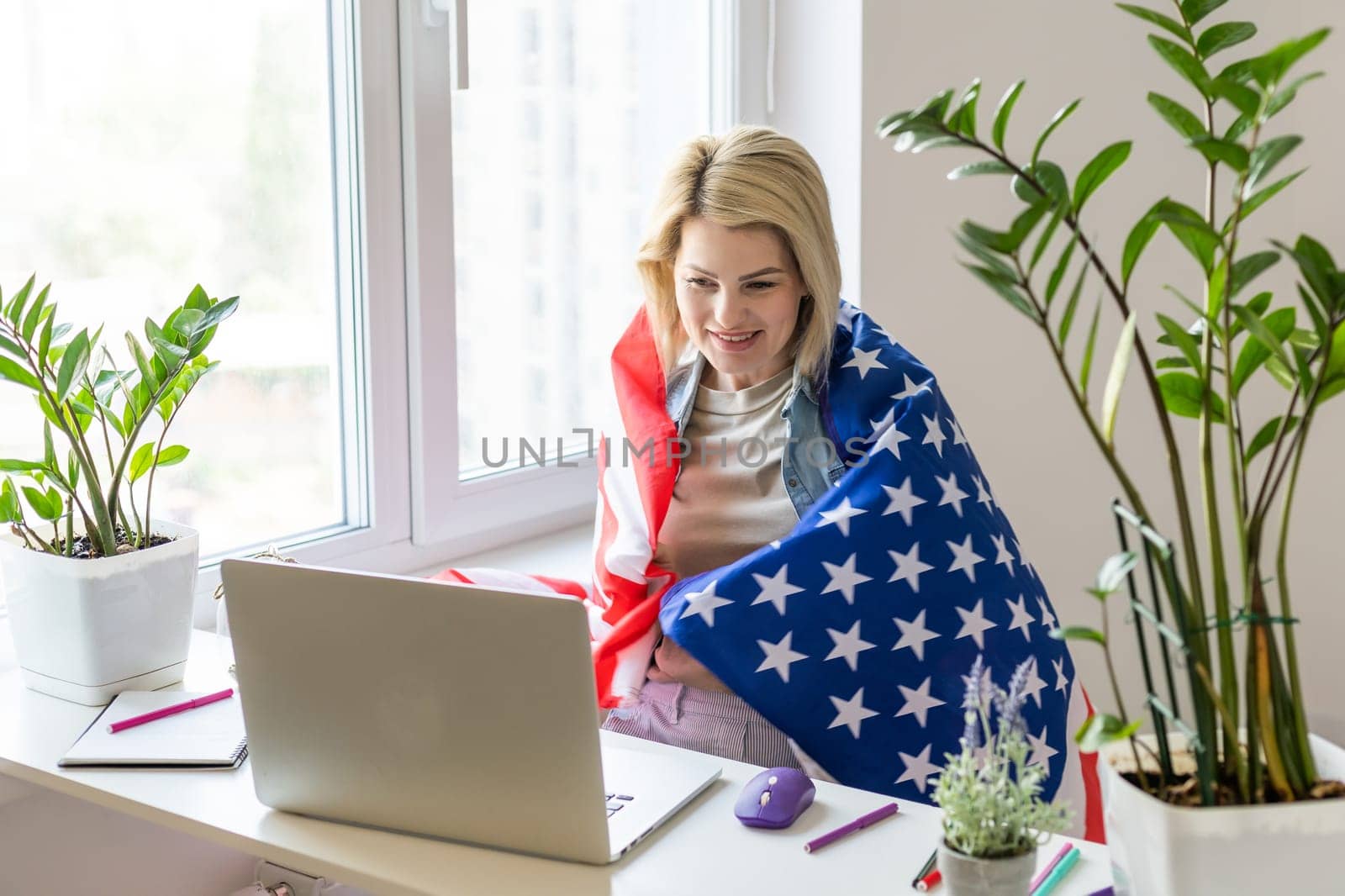 Happy woman employee sitting wrapped in USA flag, shouting for joy in office workplace, celebrating labor day or US Independence day. Indoor studio studio shot isolated on yellow background.