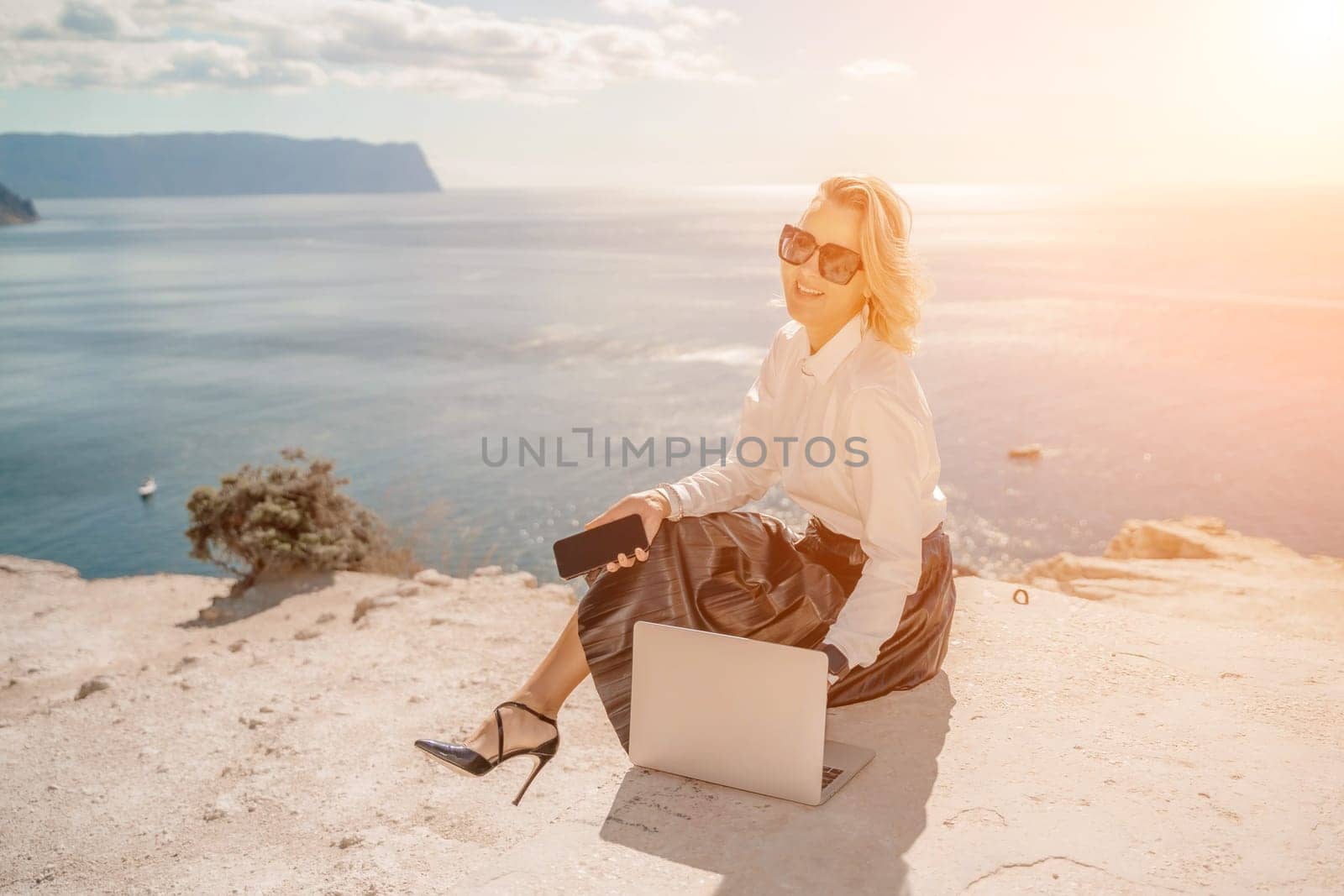 Freelance women sea working on a computer. Pretty middle aged woman with computer and phone outdoors with beautiful sea view. The concept of remote work. by Matiunina