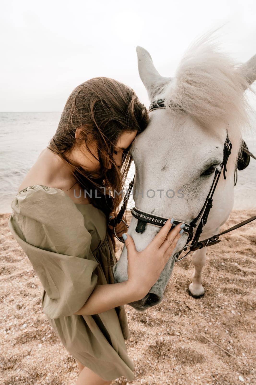 A woman in a dress stands next to a white horse on a beach, with the blue sky and sea in the background