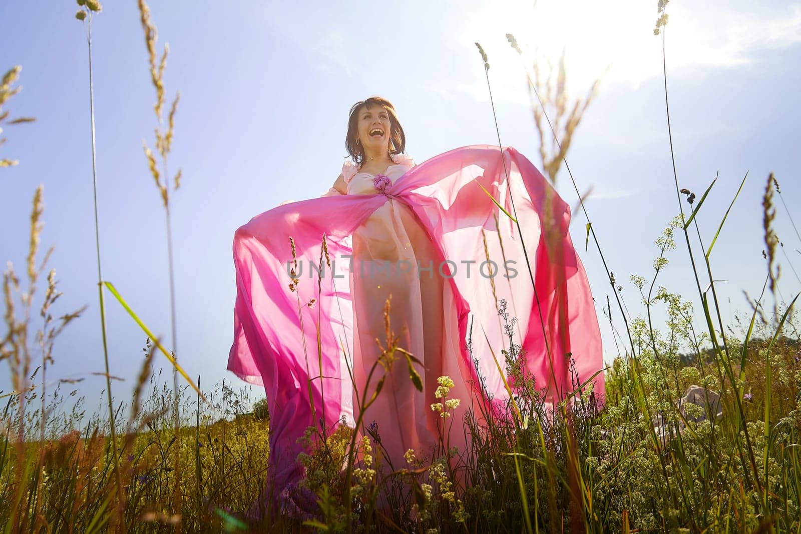 Beautiful girl in a lush pink ball gown in green field during blooming of flowers and blue sky on background. Model posing on nature landscape as princess from fary tale by keleny