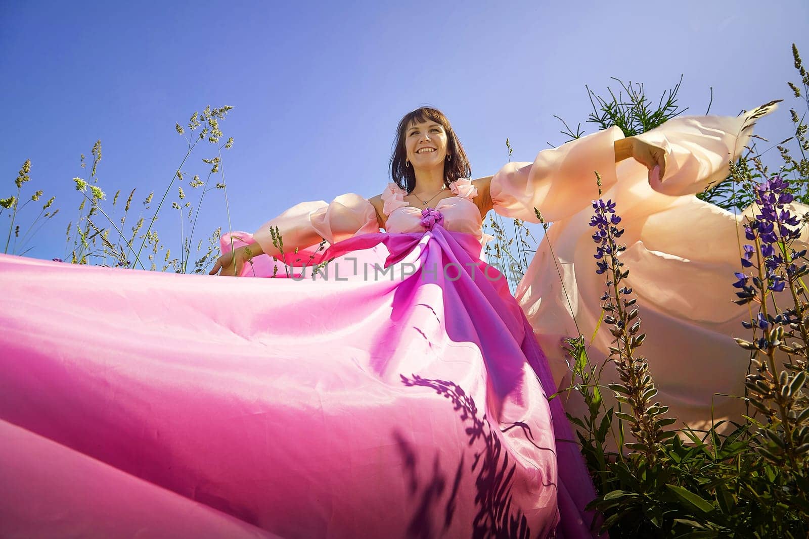 Beautiful girl in a lush pink ball gown in green field during blooming of flowers and blue sky on background. Model posing on nature landscape as princess from fary tale by keleny