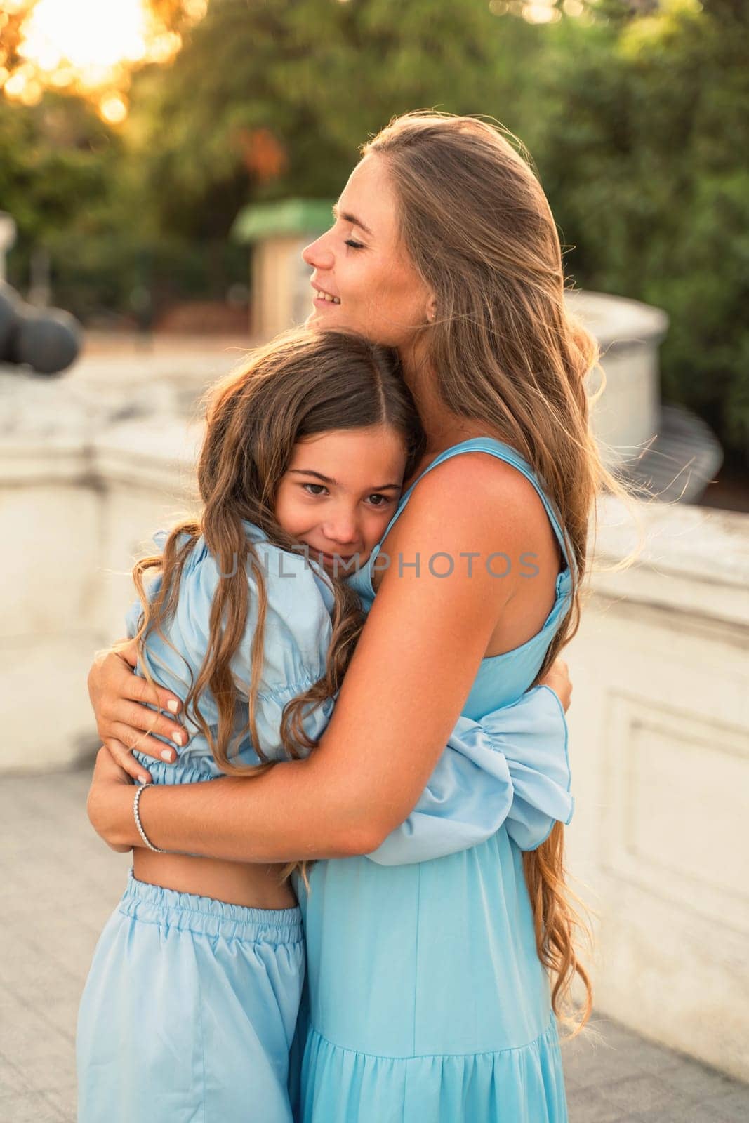 Mother aughter sunset. in blue dresses with flowing long hair against the backdrop of sunset. The woman hugs and presses the girl to her