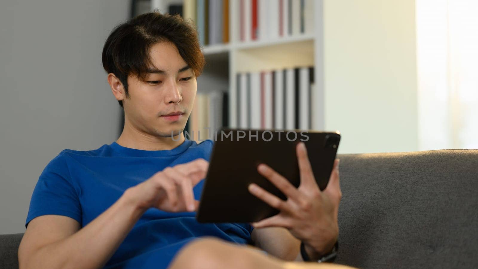 Carefree asian male freelancer sitting on sofa and checking email or working online on digital tablet.