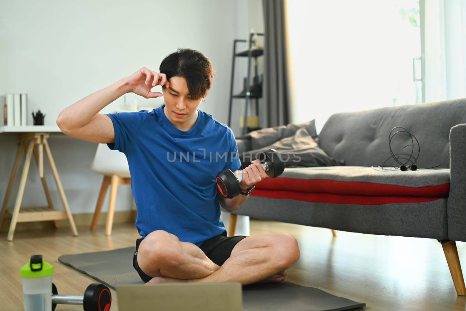 Active sportsman training arms with dumbbell and watching video online training on laptop at home by prathanchorruangsak