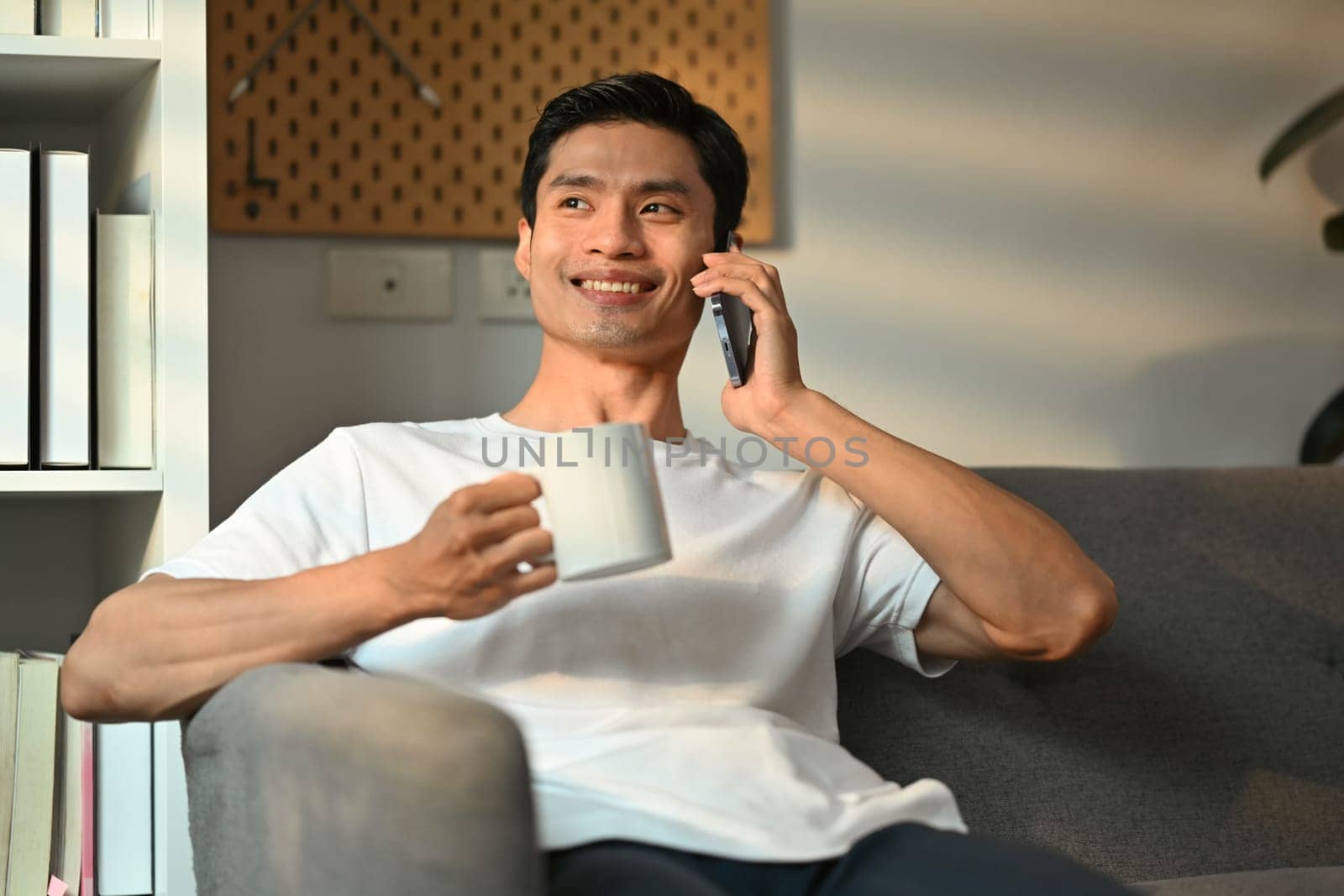 Positive millennial man having pleasant mobile phone conversation and drinking coffee in living room.