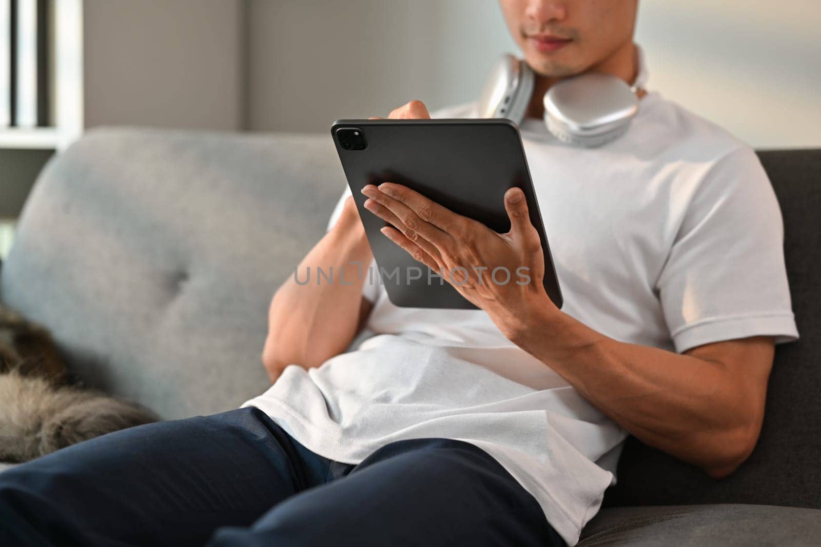 Man in casual clothes and headphone on neck using digital tablet on couch. People, technology and communication concept.