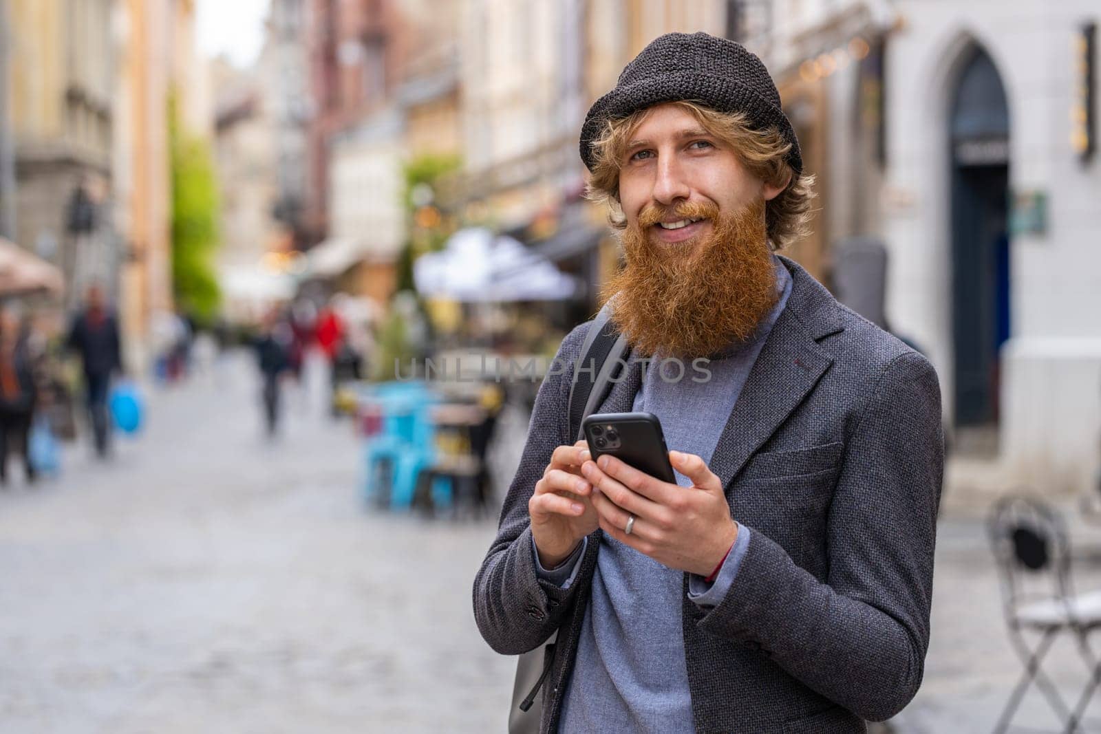 Smiling young man using smartphone typing text messages browsing internet, finishing work, looking at camera. Redhead bearded guy walking in urban city street background. Town lifestyles outdoors