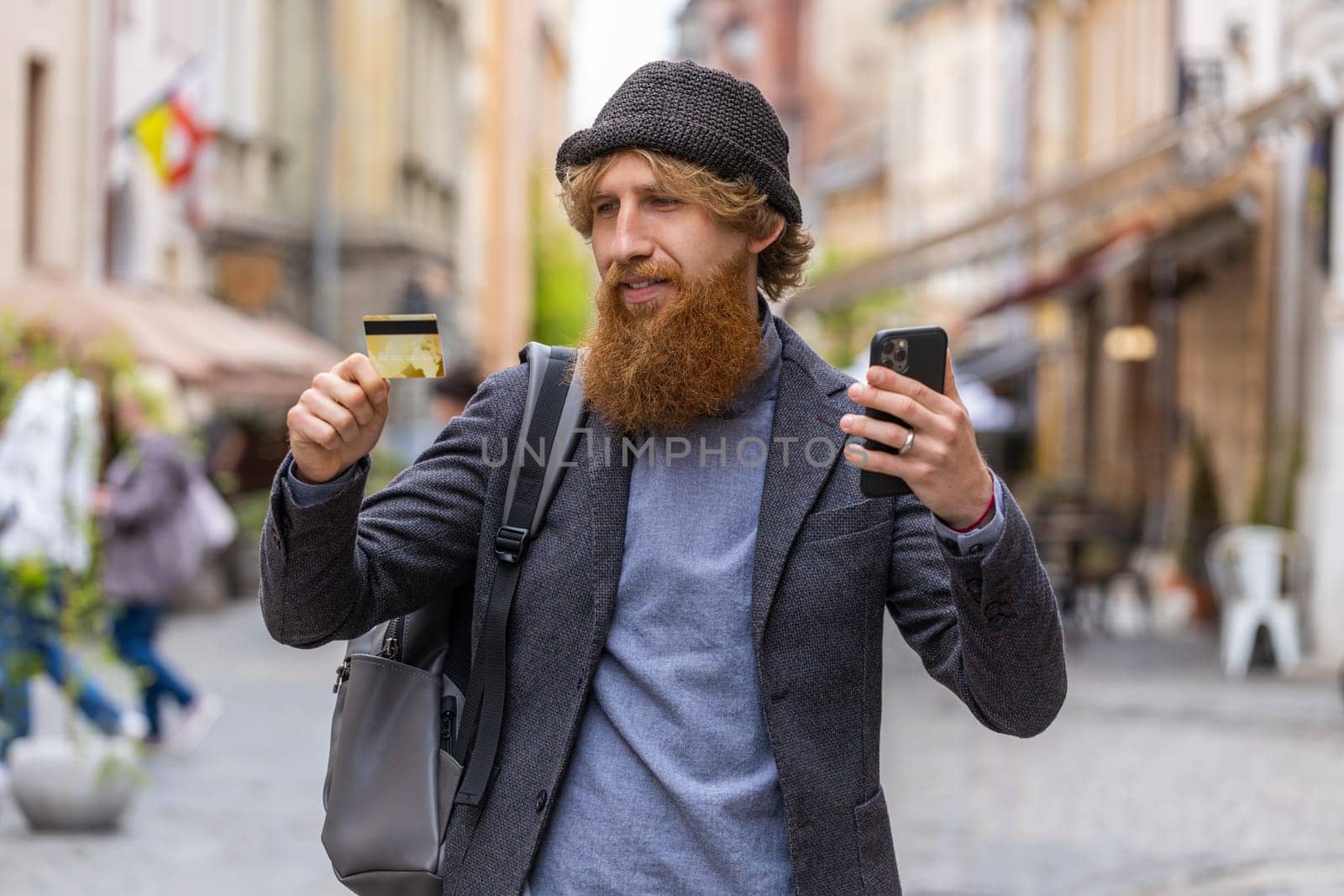 Young bearded man using credit bank card smartphone while transferring money, purchases online shopping, order food delivery, booking hotel room. Redhead guy walking in urban city street outdoors
