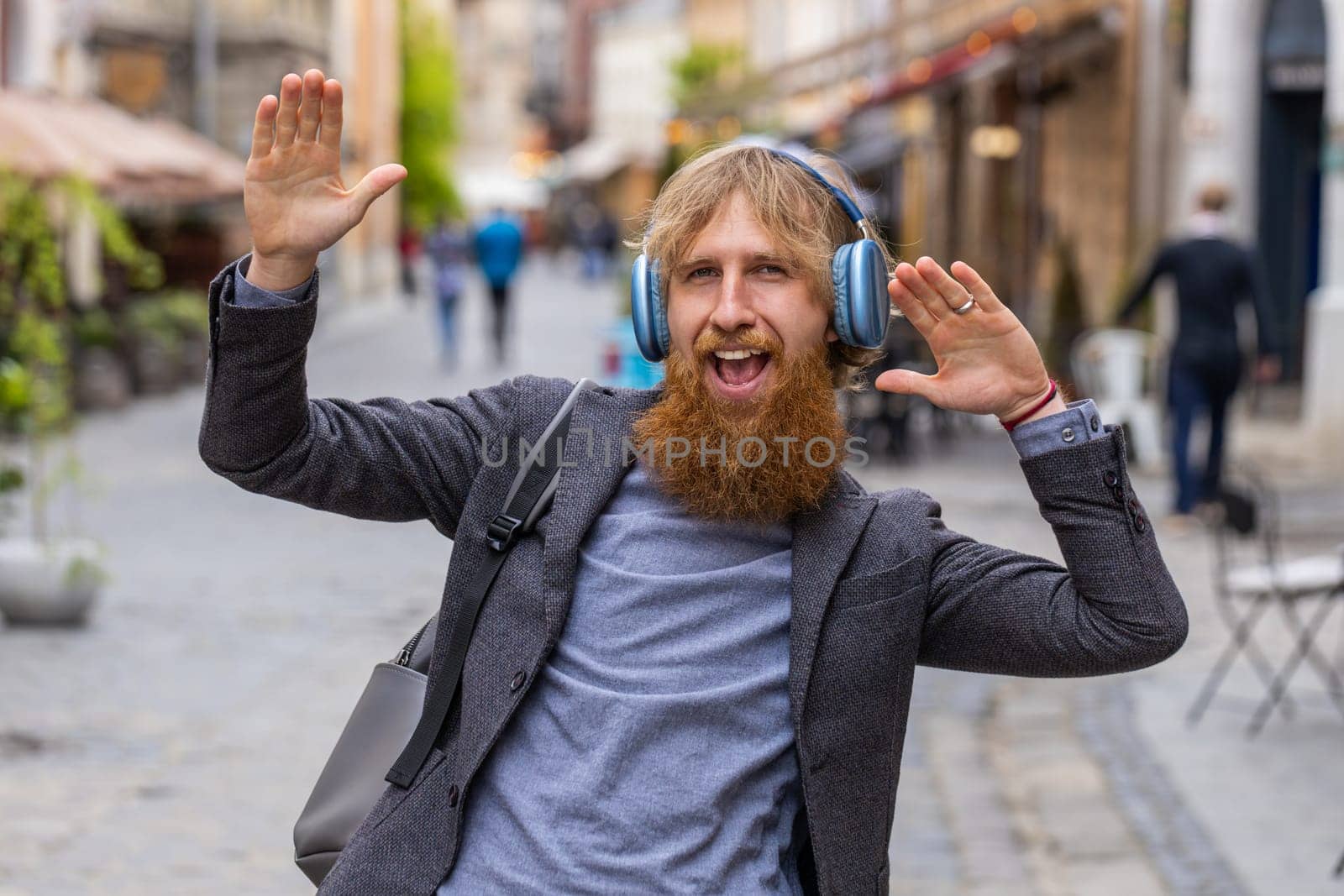 Happy overjoyed young man in wireless headphones listening favorite energetic music celebrating win. Redhead bearded guy tourist traveler walking in urban city street. Town lifestyles outdoors weekend