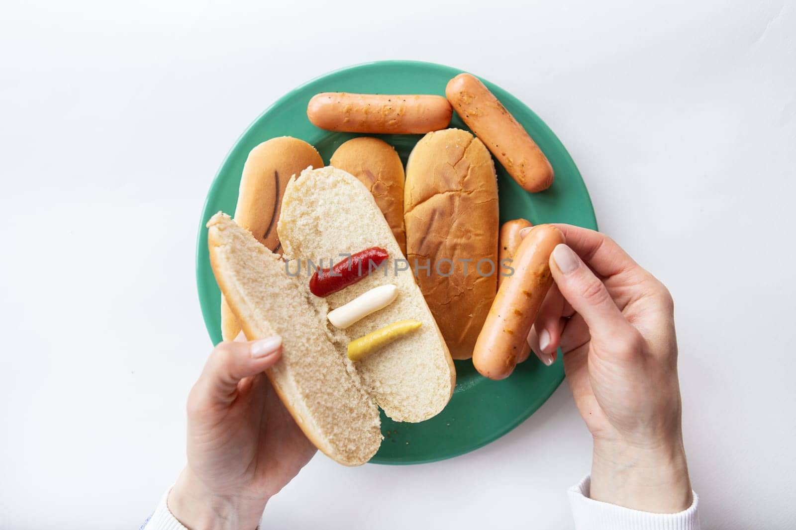 Fresh hot dog buns which lie on a green plate along with sausages. The girl holds in her hands a bun with sauces in which she puts a sausage. by sfinks