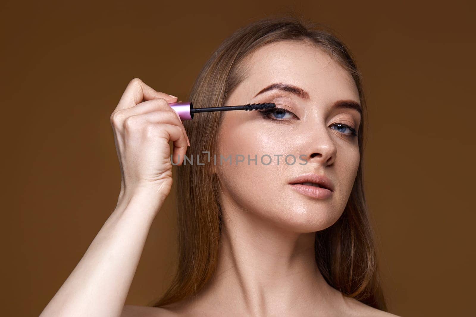 Beautiful woman applying black mascara on long thick eyelashes with brush on beige background
