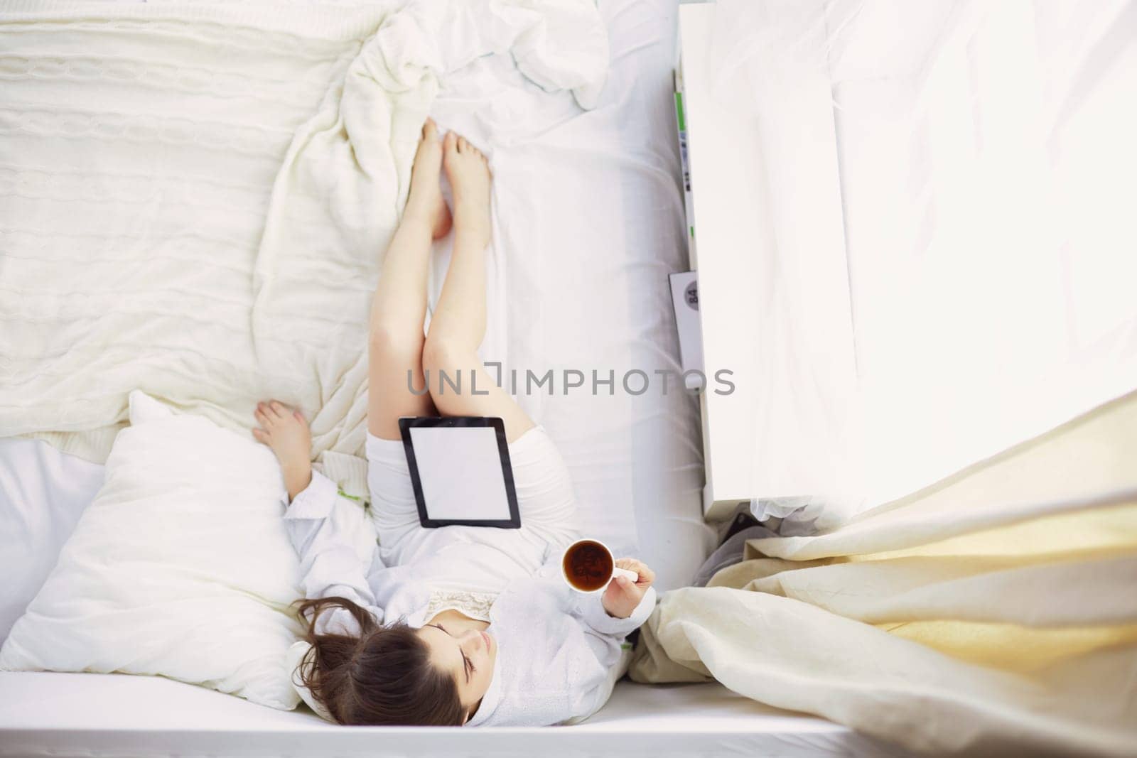 Portrait of woman using tablet and drinking coffee while sitting on the bed in the morning.