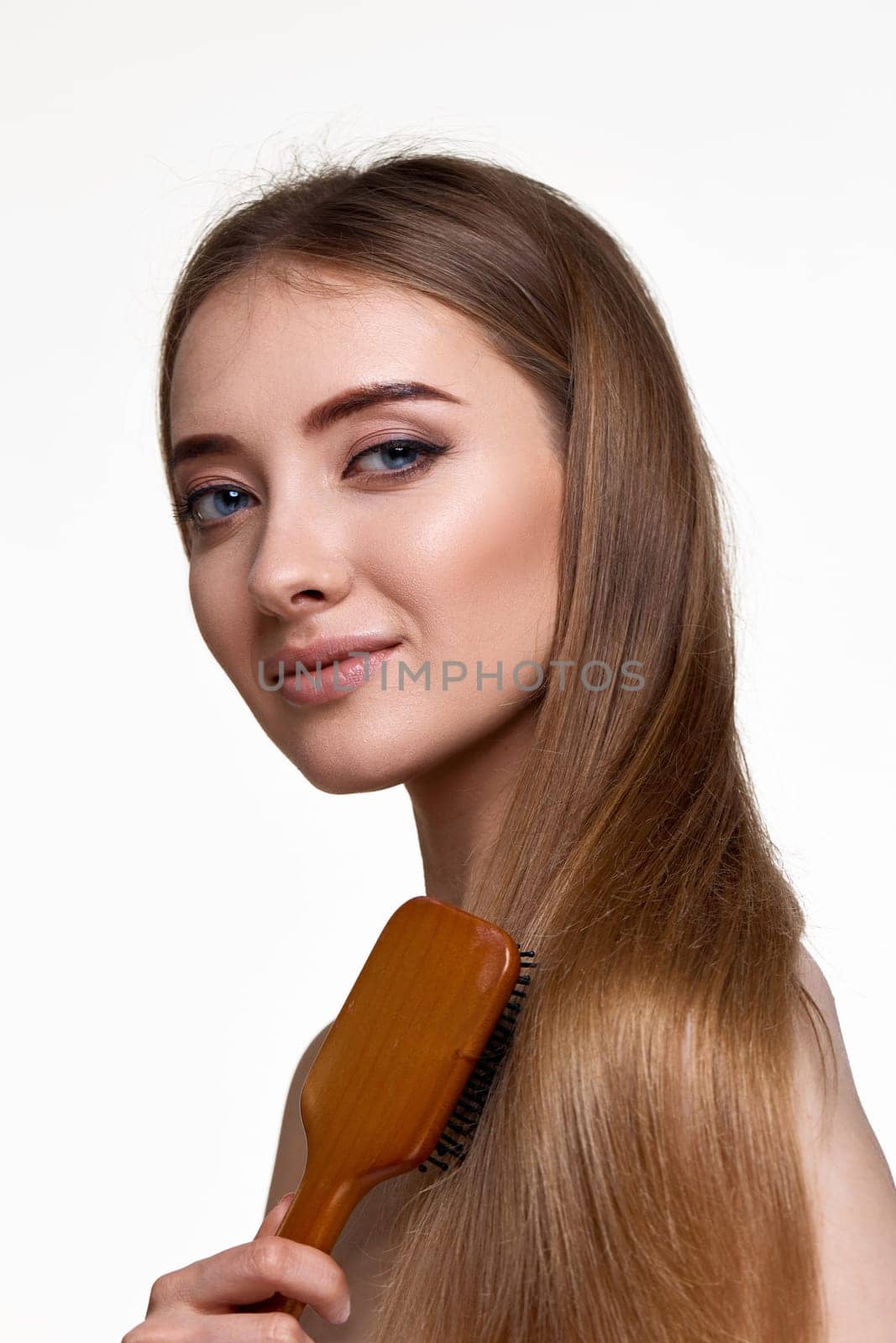 Beautiful smiling brunette woman looks at the camera and combs her hair with wooden brush isolated on white background