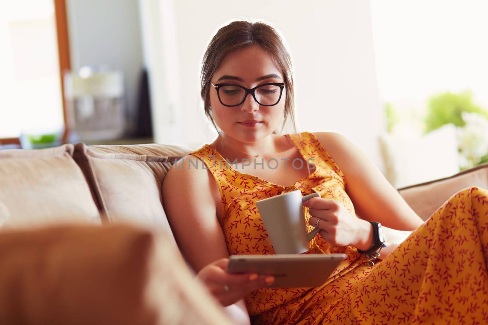 Young woman uses tablet while relaxing at home with coffee on the sofa couch.