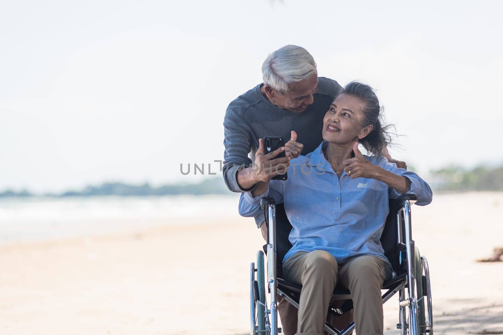 elderly woman sitting in wheelchair and husband is a wheelchair user smartphone taking selfie on the beach by Sorapop