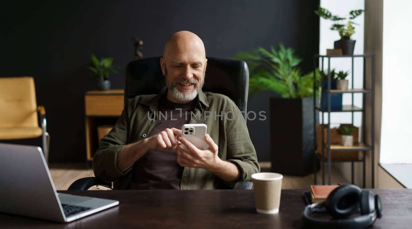 Bald and respectable man with gray beard. He captured in moment of joy, smiles while reading messages on phone. Computer, cup of coffee, and audio headphones are present on the table. by LipikStockMedia