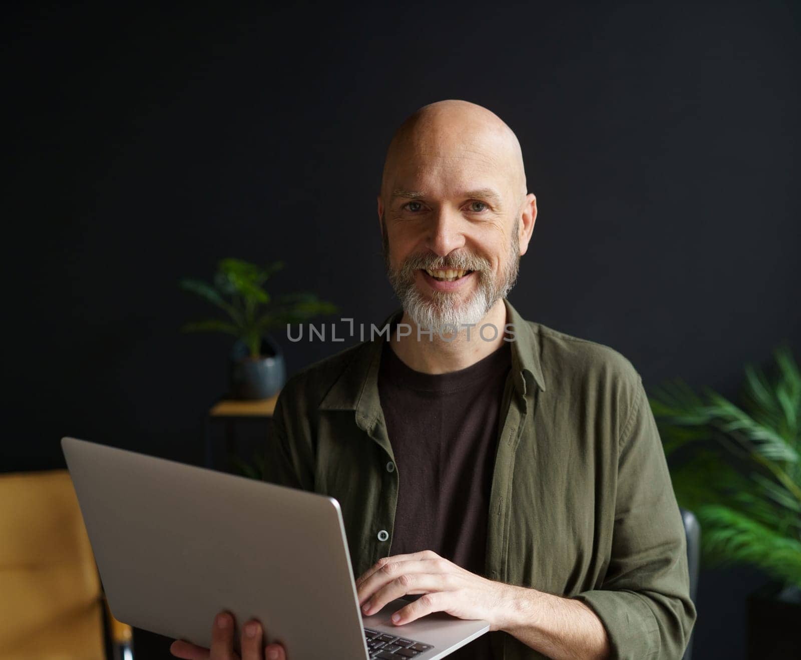 Happy senior man with warm smile, holding laptop in hands home. Man's cheerful and joyful expression, highlighting his contentment and happiness. by LipikStockMedia